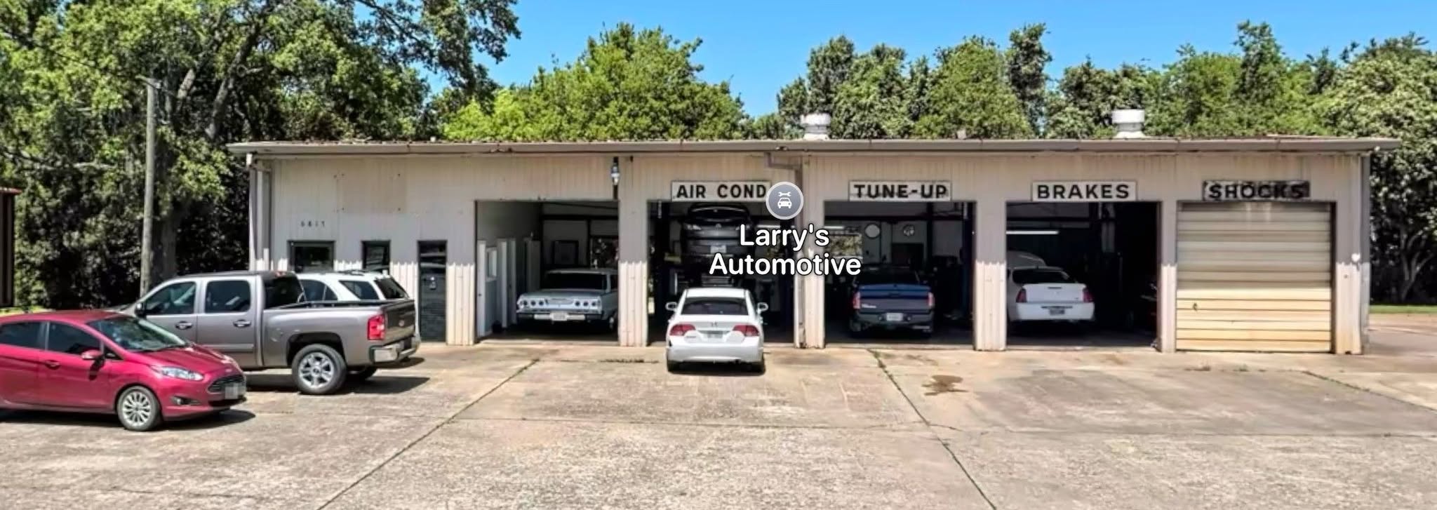 The beige metal shop at 6817 Bayway Drive with open bays marked AIR COND, TUNE-UP, BRAKES, and SHOCKS, and cars on the concrete lot.