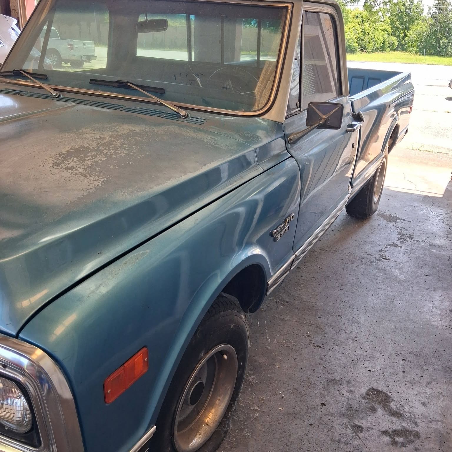 A teal Chevrolet C10 in the shop on stained concrete, open bay and trees behind it.